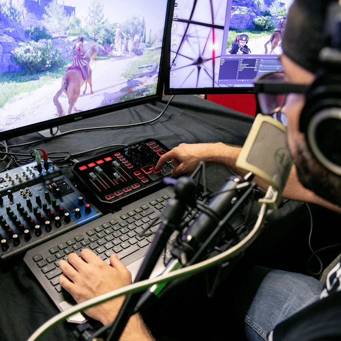 A streaming desk: GoXLR beside a mixing console, keyboard, microphone on a boom arm and two monitors running a game and a capture app.